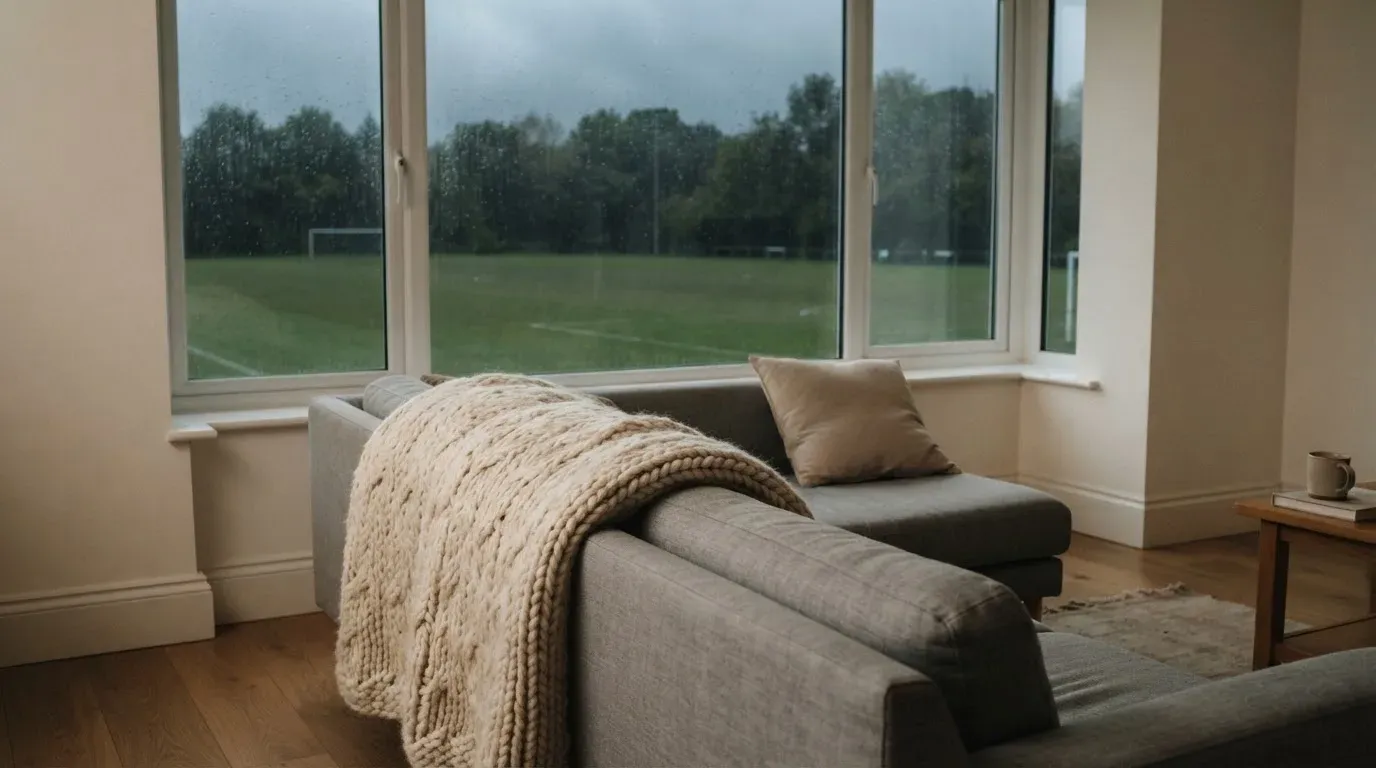 Ein Sofa mit Wolldecke steht vor einem grossen Fenster, draussen Regen und in der Ferne ein Fussballplatz.