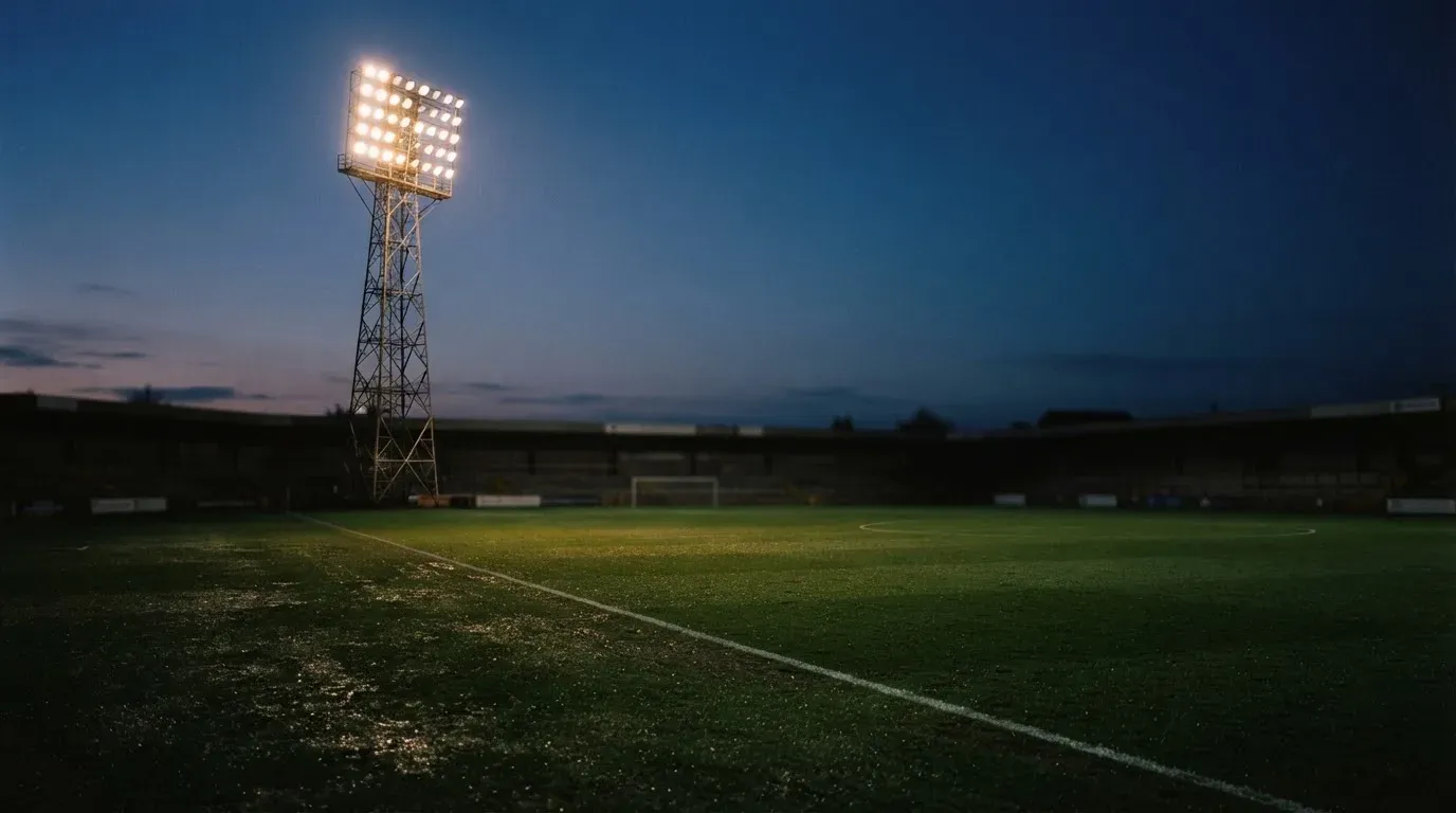 Ein Flutlichtmast ragt in den Abendhimmel, die Rasenfläche darunter liegt leer und dunkelgrün da.