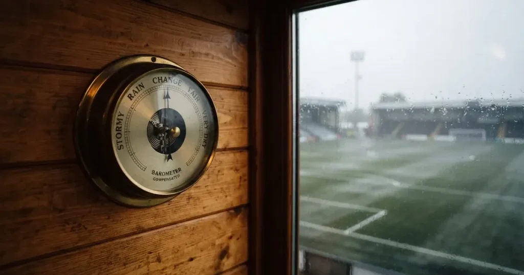 Ein Wetterbarometer hängt an einer Holzwand neben einem Fenster, draussen liegt ein nasser Fussballplatz im Regen.