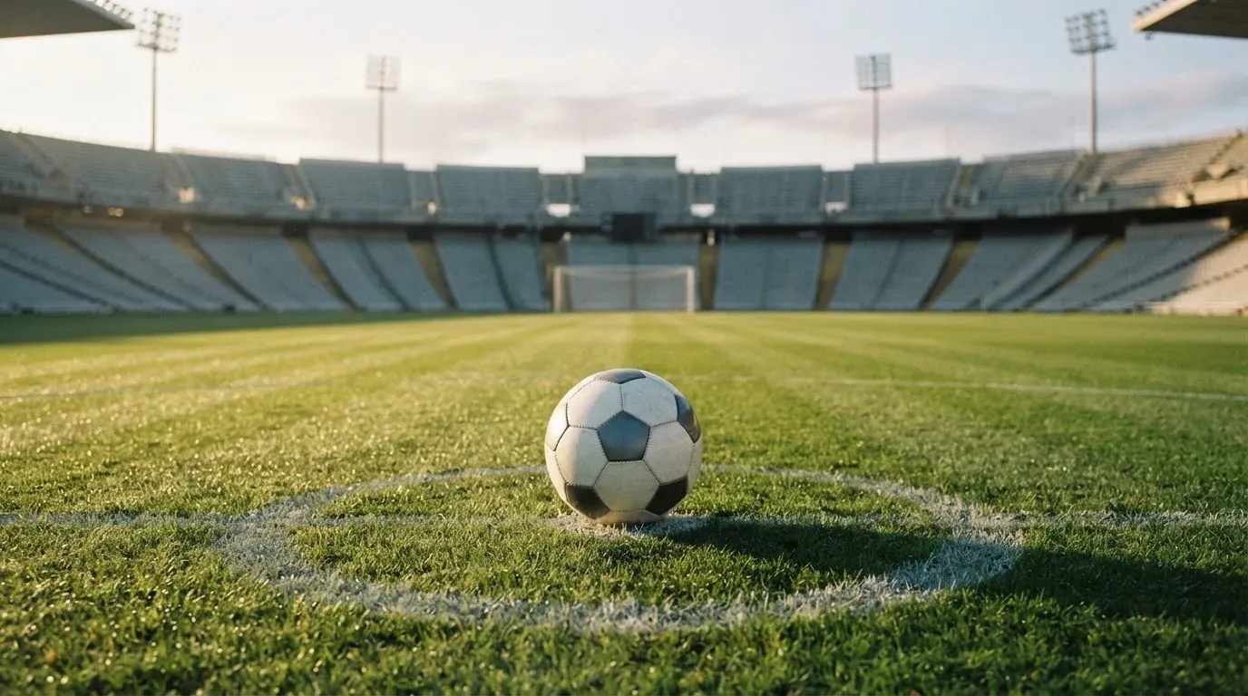 Ein Fussball liegt auf dem Anstosspunkt eines leeren Rasenplatzes im frühen Morgenlicht, dahinter erhebt sich eine menschenleere Tribüne.