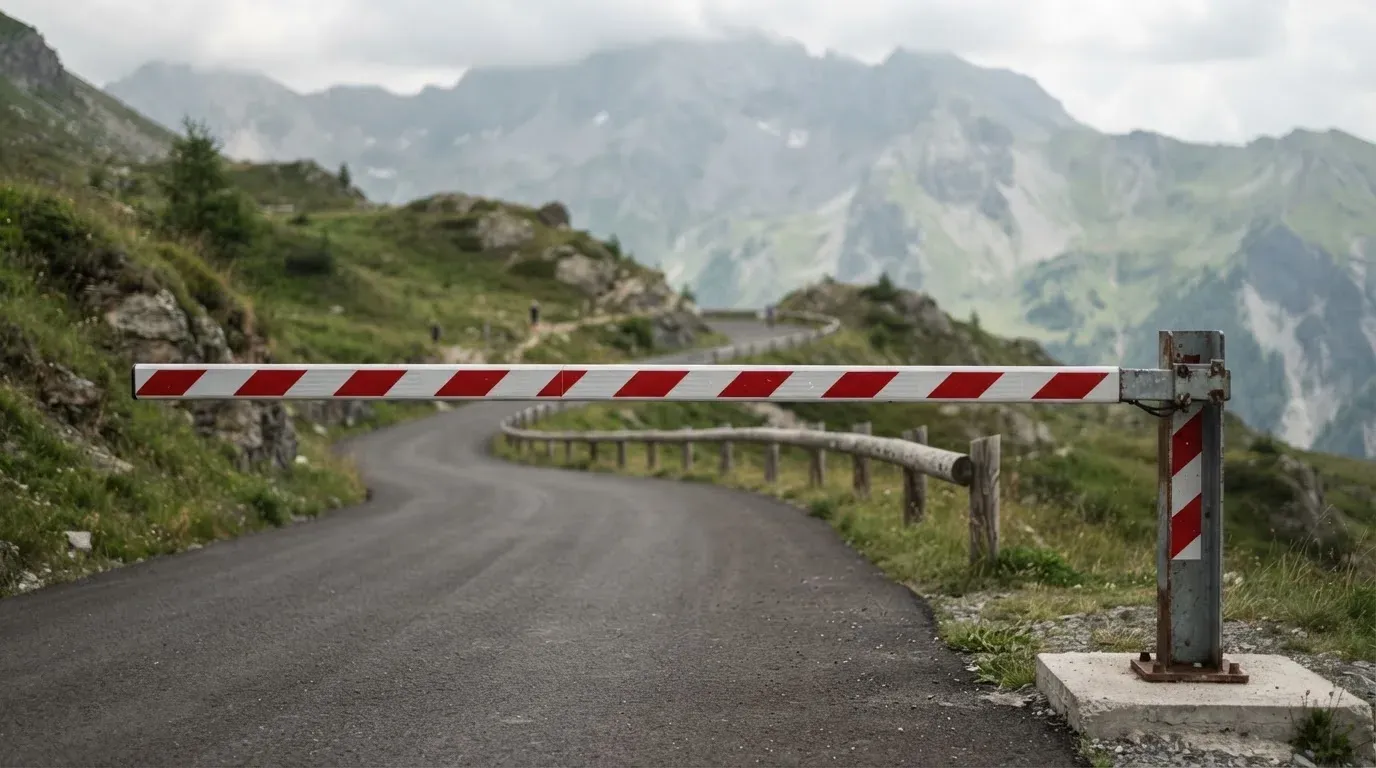 Ein Schlagbaum sperrt eine Landstrasse in den Bergen, dahinter windet sich der Weg zwischen grünen Hängen.
