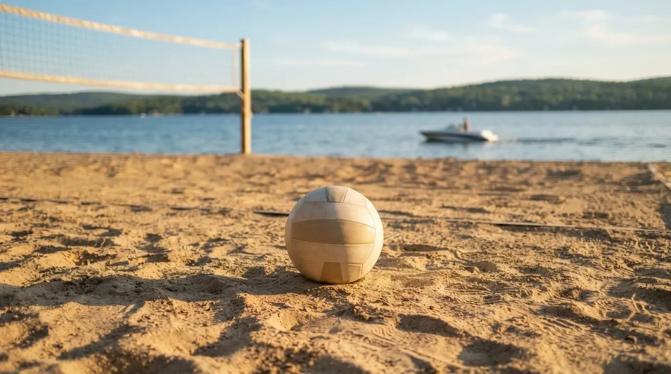 Ein Volleyball liegt im Sand eines Spielfelds am Seeufer, im Hintergrund gleitet ein Boot über das Wasser.