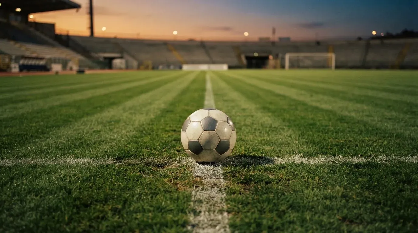 Ein Fussball liegt auf dem Mittelkreis eines leeren Rasenplatzes, dahinter ragen leere Tribünenreihen in den Abendhimmel.
