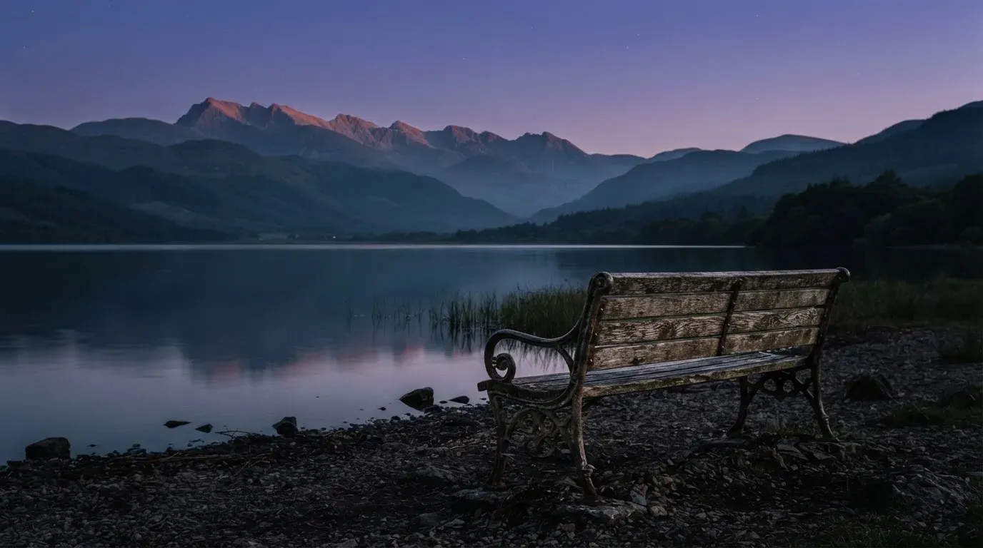Eine Holzbank steht in der Dämmerung am Seeufer, im Hintergrund zeichnet sich ein Bergpanorama gegen den violetten Himmel ab.