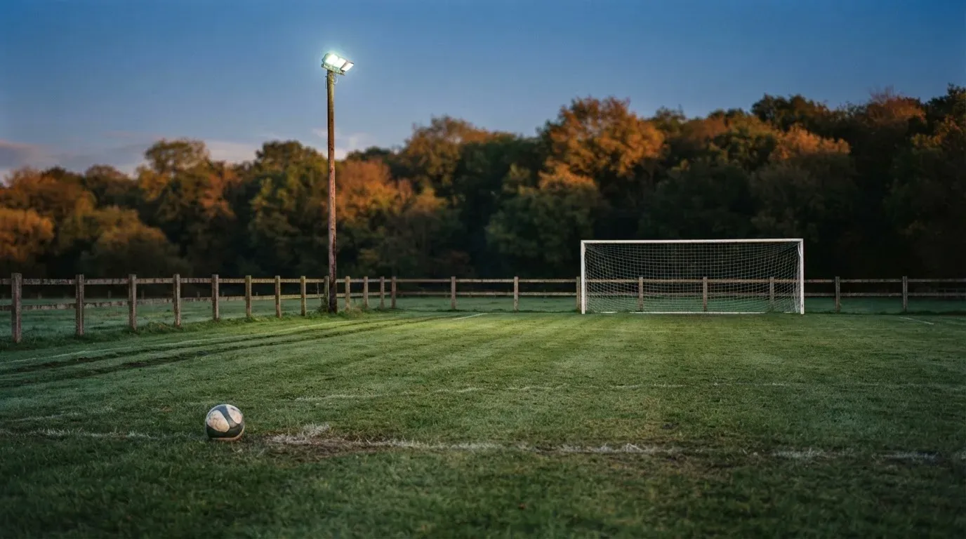 Ein kleiner Dorf-Fussballplatz liegt in der Dämmerung unter einem hohen Flutlichtmast, am Spielfeldrand steht ein Holzzaun.