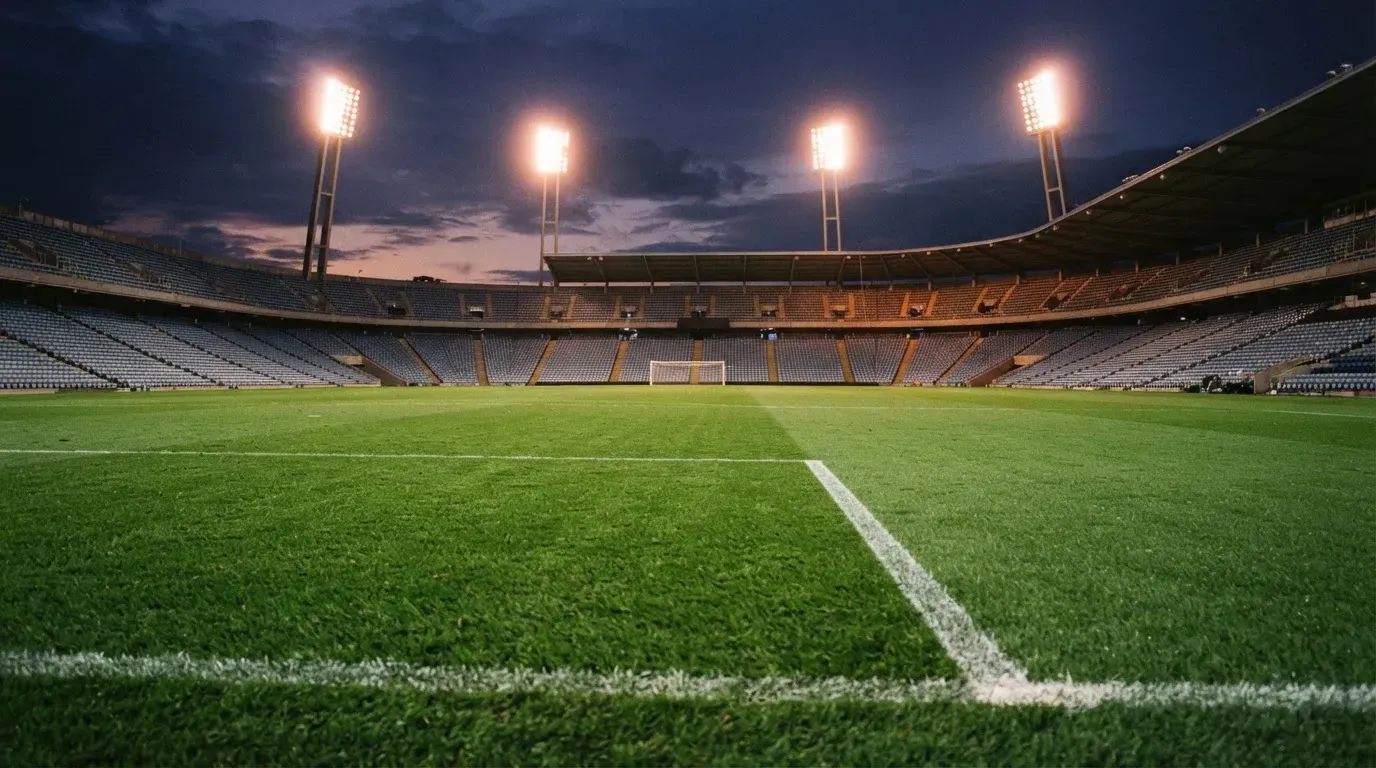 Leere Tribünenränge eines Fussballstadions in der Abenddämmerung, über dem Rasen leuchten die Flutlichter.