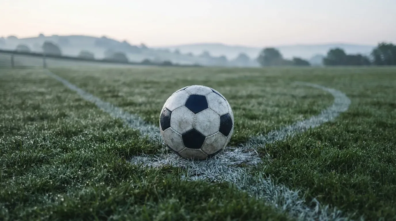 Ein Fussball liegt am frühen Morgen auf dem Anstosspunkt eines taunassen Rasens, im Hintergrund erheben sich Hügel im Dunst.