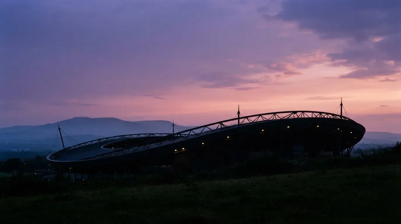 Die Silhouette eines Stadiondachs vor dem violetten Abendhimmel, dahinter eine Hügelkette.