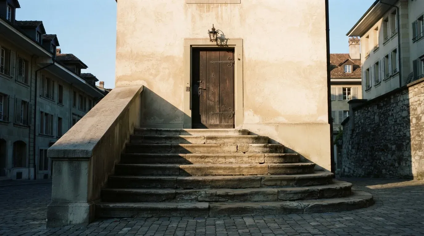Eine Steintreppe führt zu einer schweren Eichentür in einer hellen Fassade, die Stufen liegen im Morgenschatten.