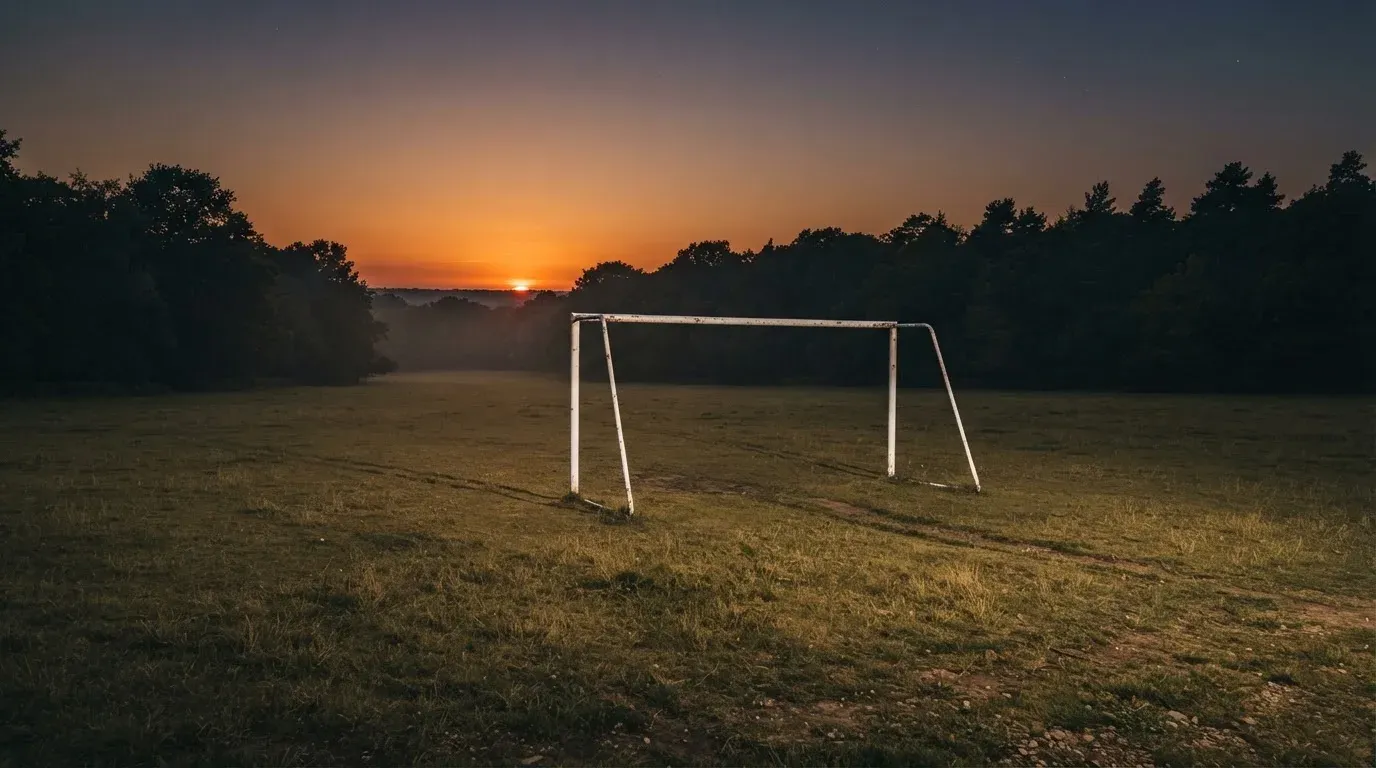 Ein Fussballtor ohne Netz steht auf einer Wiese am Waldrand, die Abenddämmerung färbt den Himmel orange.
