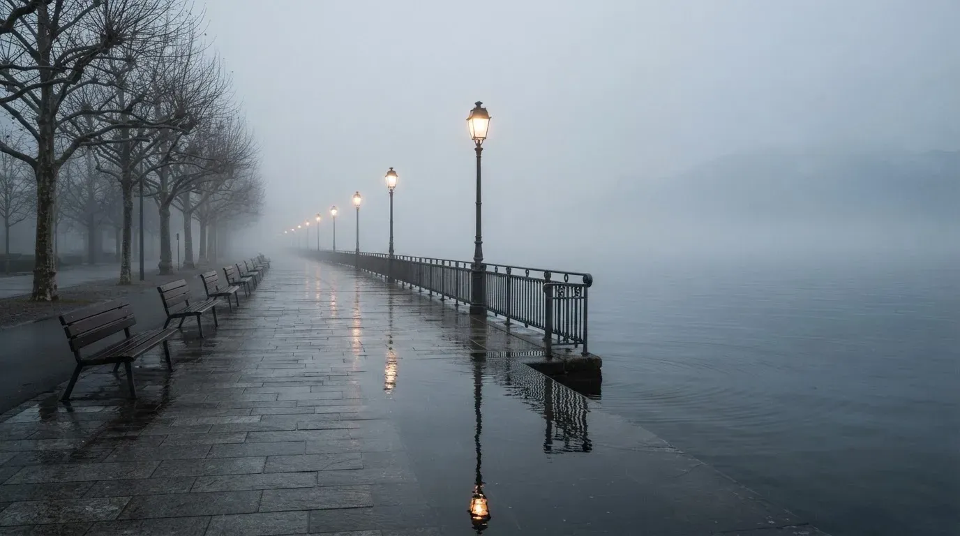 Eine Seepromenade mit leeren Bänken an einem nebligen Morgen, im Wasser spiegeln sich die Lichter der Uferlaternen.