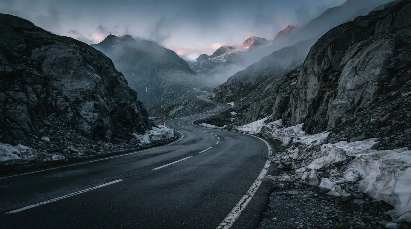 Ein Bergpass in den Schweizer Alpen mit leerer Strasse und Schneeresten am Rand im ersten Frühlicht.