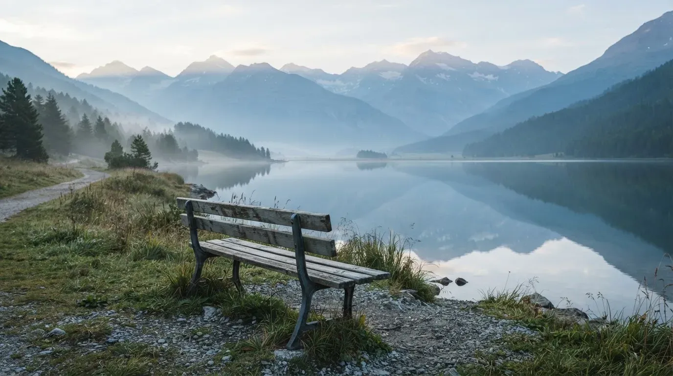 Eine Holzbank an einem Seeufer mit Blick auf eine Bergkette im Morgendunst, das Wasser ist spiegelglatt.