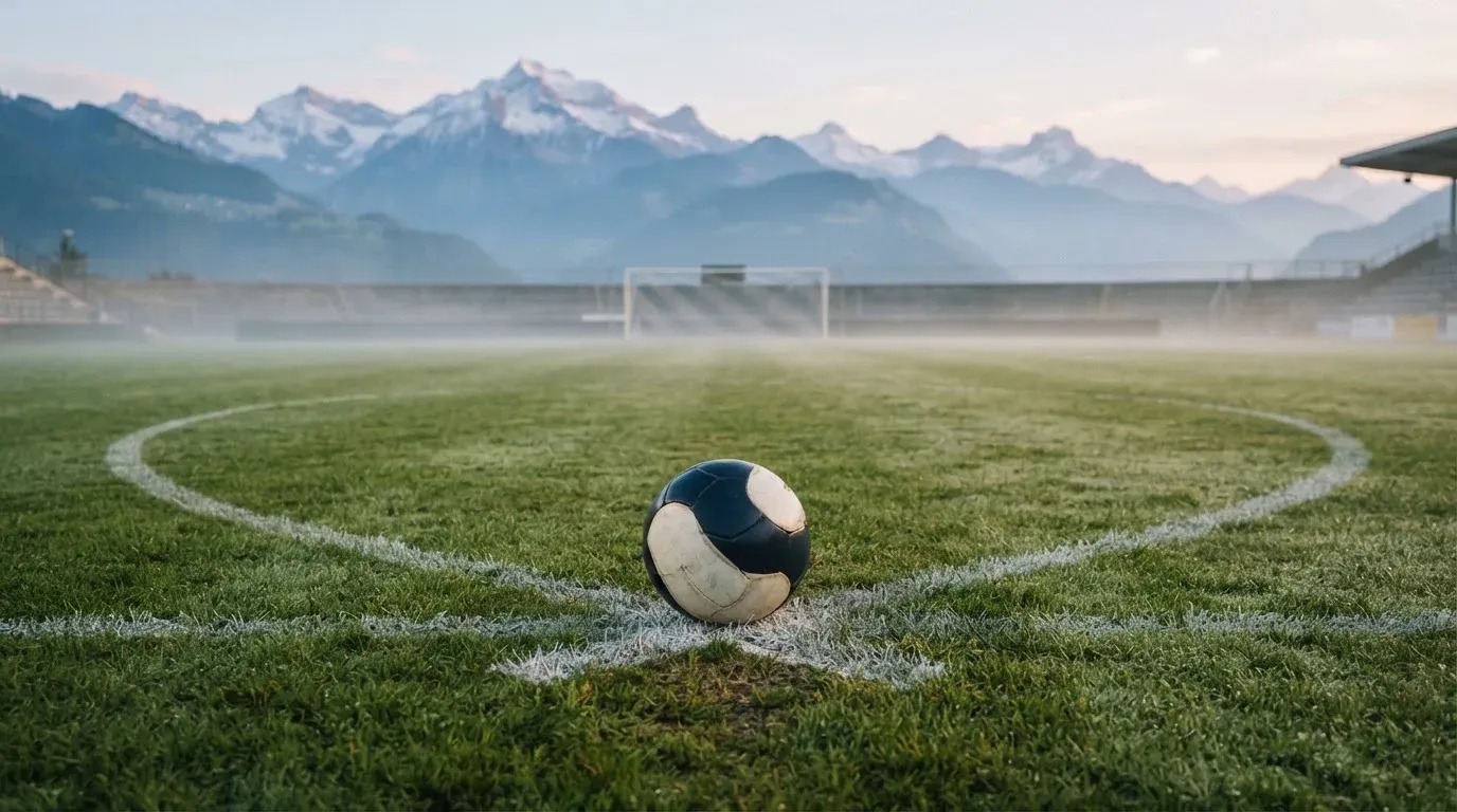 Ein einzelner Fussball auf dem Mittelpunkt eines leeren Rasens im Morgennebel, hinter dem Tor ragen Berge auf