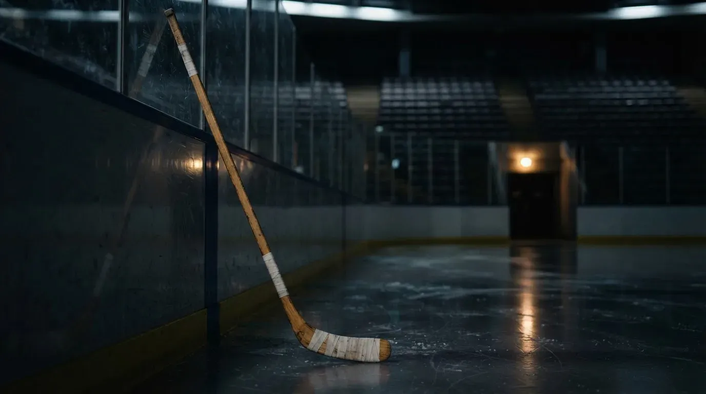 Ein Eishockeyschläger lehnt an einer Bande, im Hintergrund eine fast leere Tribüne im gedimmten Hallenlicht