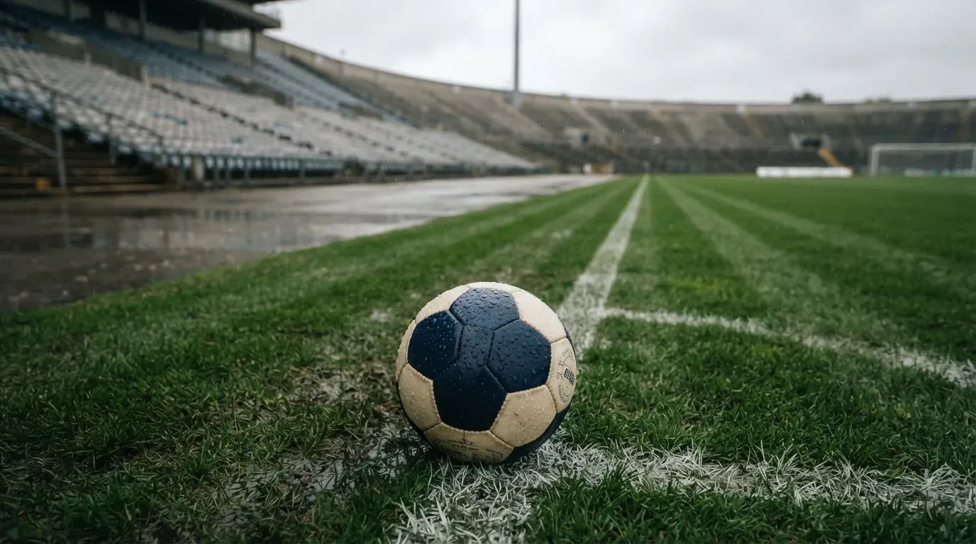 Ein Fussball liegt auf dem Anstosspunkt eines nassen Rasens, dahinter eine leere Tribüne unter grauem Himmel.