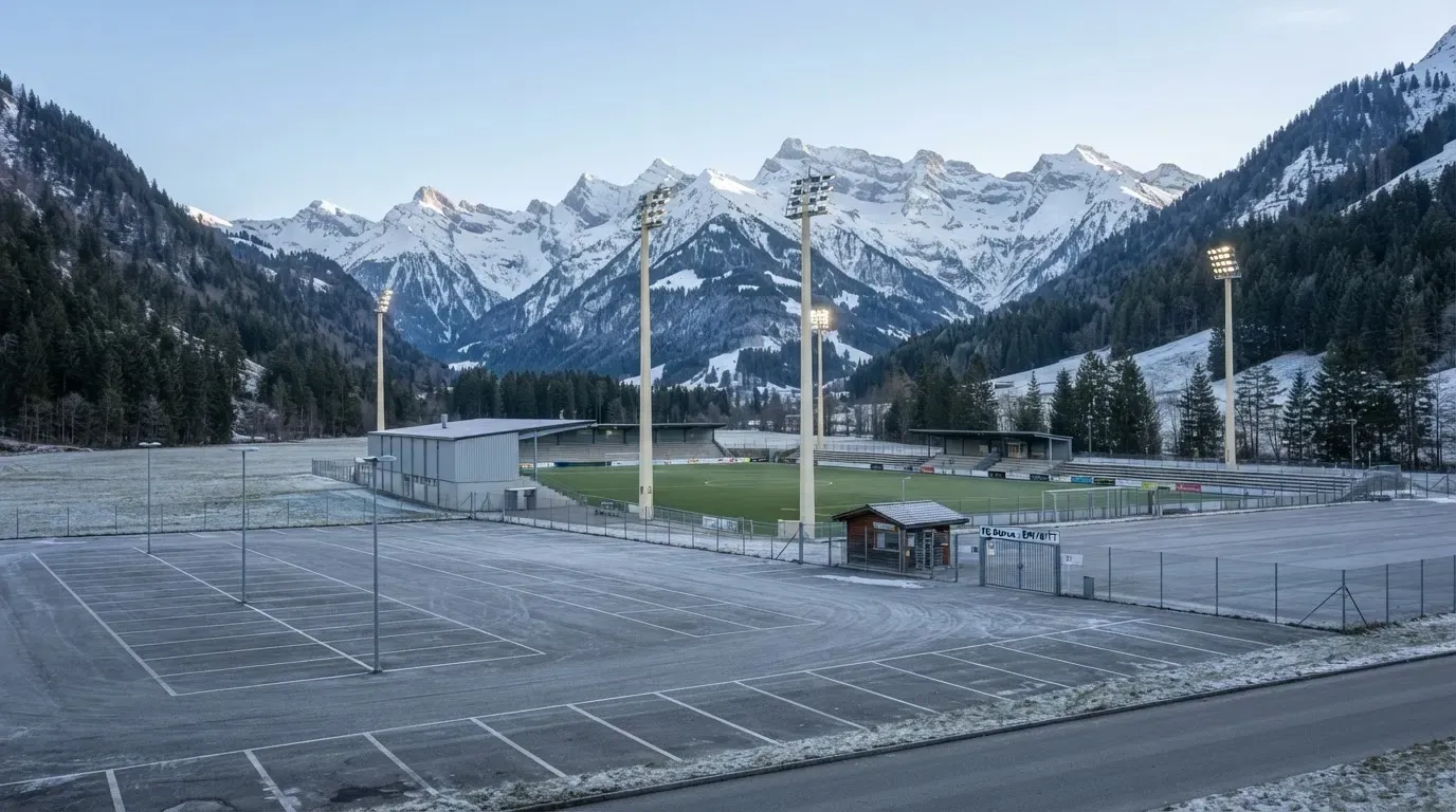 Schneebedeckte Berge ragen hinter einem kleinen Stadion in einem Alpental auf, im Vordergrund ein leerer Parkplatz am Morgen.