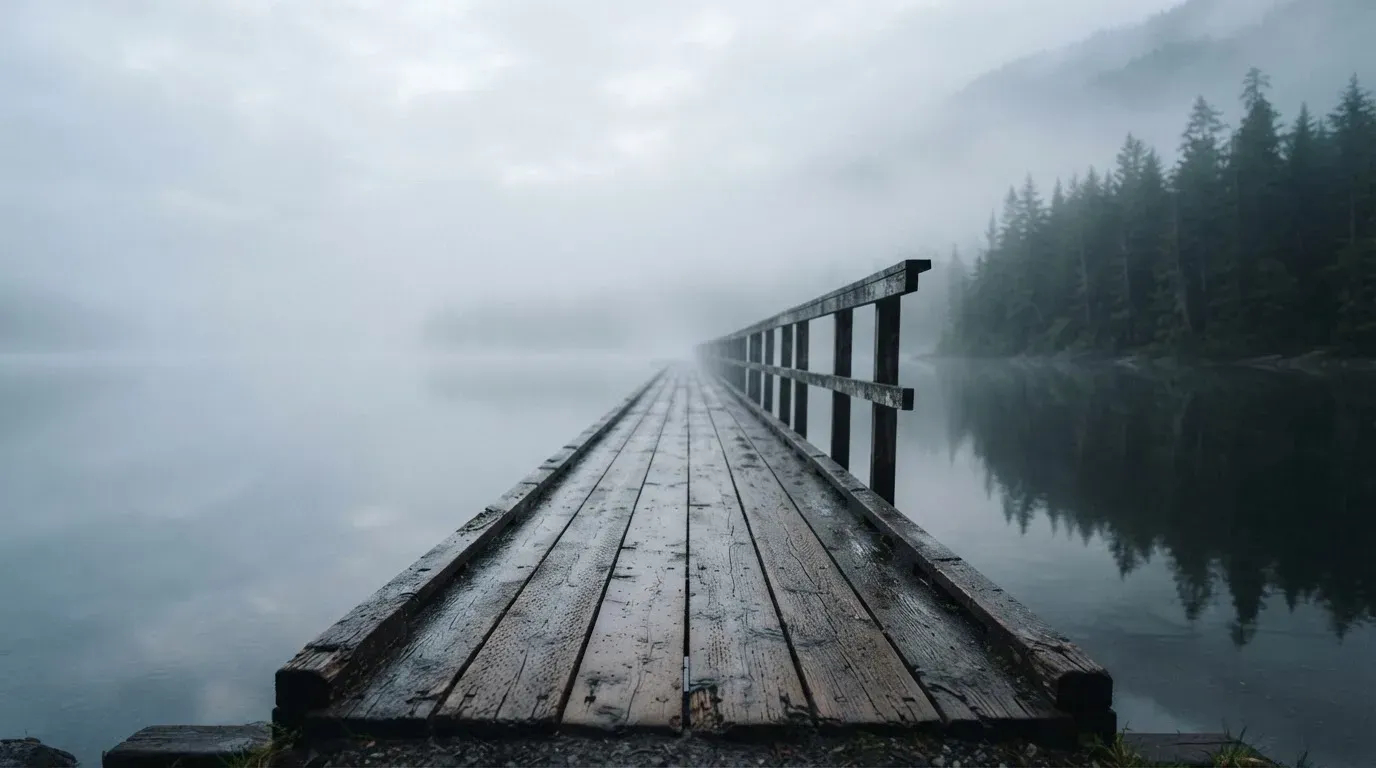 Ein Holzsteg führt über einen ruhigen Bergsee im Nebel, am anderen Ufer stehen dunkle Tannen.