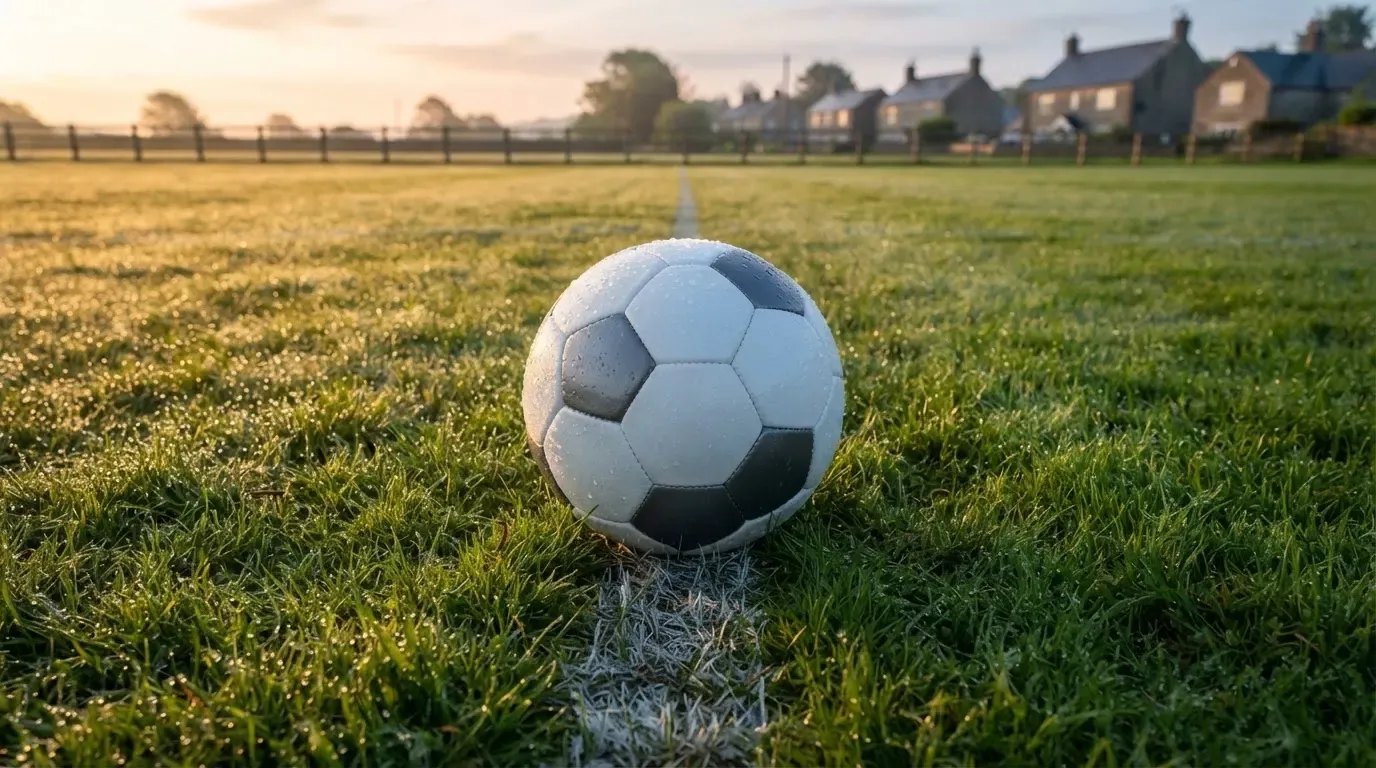 Ein Fussball liegt im Tau auf der Mittellinie eines Dorfplatzes bei Sonnenaufgang, der Rasen glitzert.