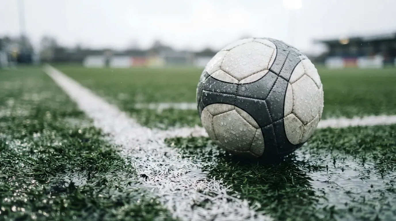 Ein Fussball liegt auf nassem Kunstrasen neben einer weissen Linie, Pfützen spiegeln den Himmel.