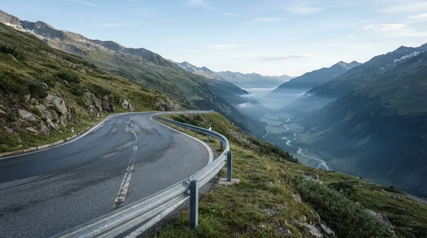 Eine Passstrasse in den Alpen mit enger Kurve und Leitplanke, darüber ein klarer Morgenhimmel und einzelne Nebelschwaden im Tal.