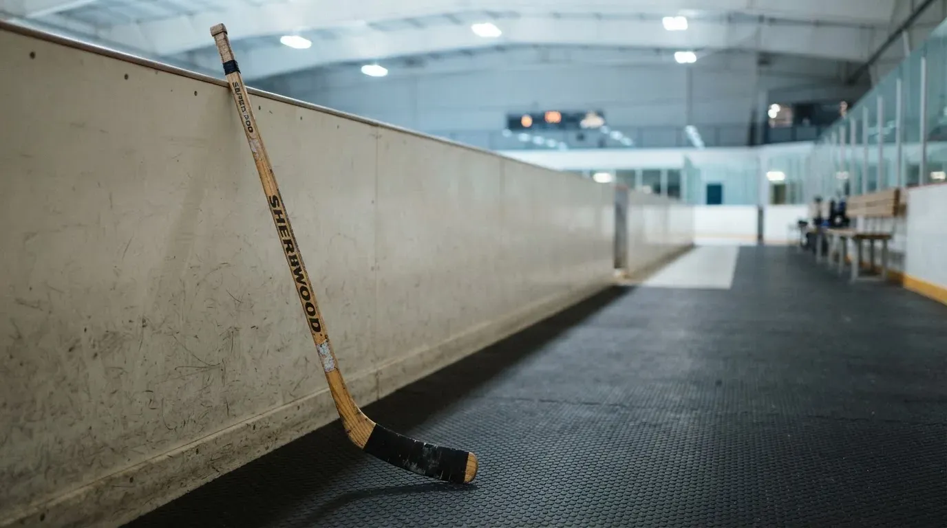 Ein Eishockeyschläger lehnt an der Bande, in der Ferne liegt die hell beleuchtete Eisfläche einer Halle unter Flutlicht.