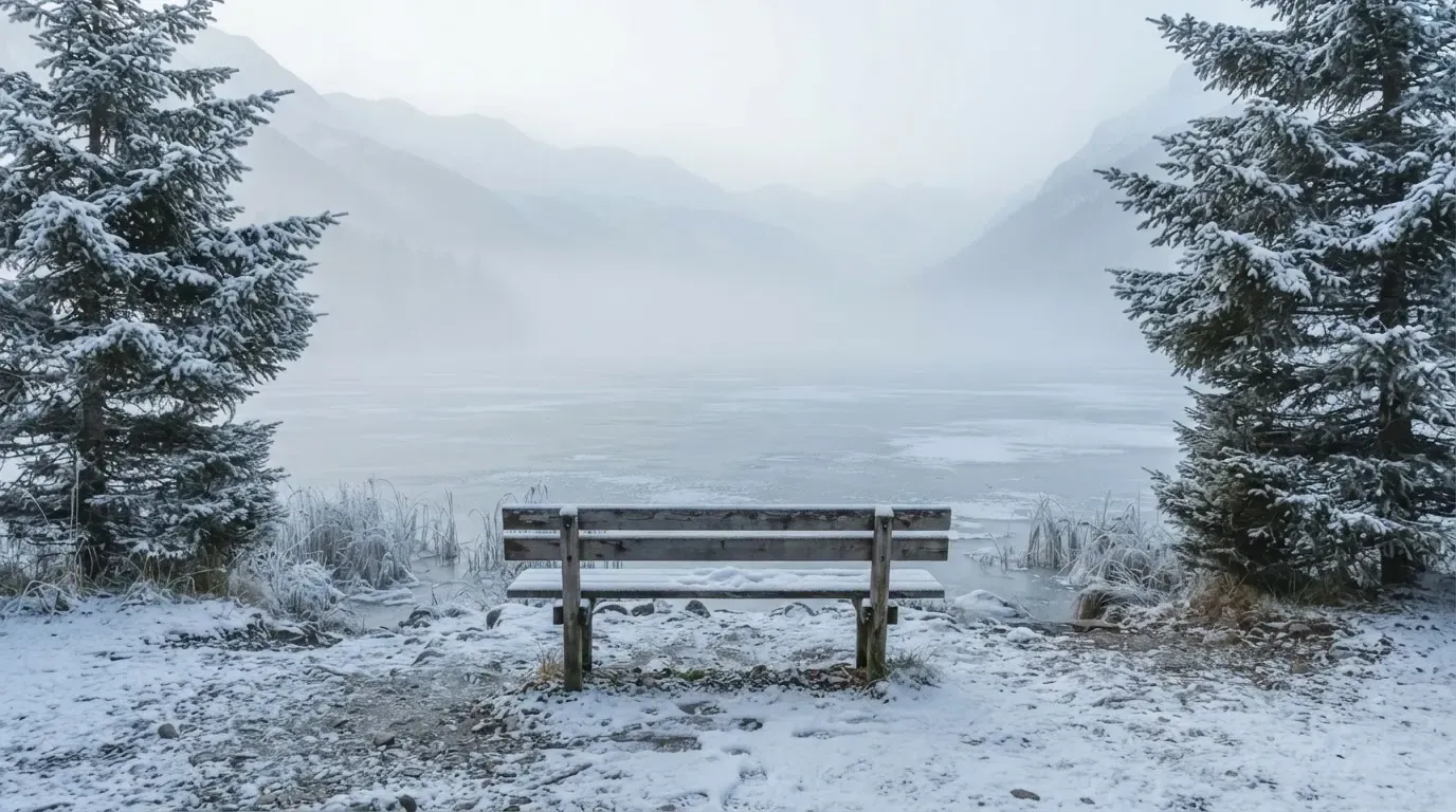 Ein zugefrorener Bergsee im Morgendunst, am Ufer steht eine einsame Holzbank zwischen verschneiten Tannen.