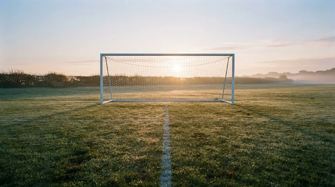 Ein Fussballtor mit Netz in der Totalen auf einem leeren Platz, Tau glänzt im Gras und die Morgensonne steht tief.