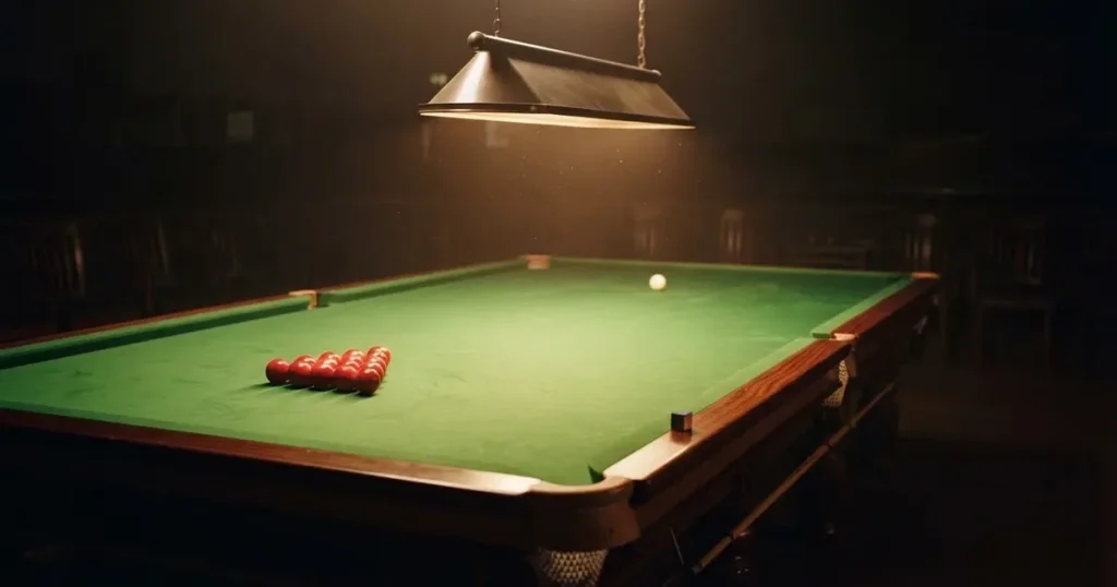 Ein Snookertisch mit grünem Tuch und einem Dreieck aus roten Kugeln unter einer tief hängenden Lampe in einer leeren Halle.