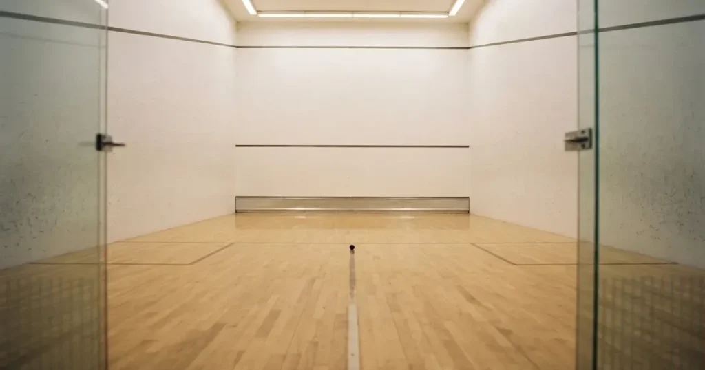 Ein leerer Squashcourt mit weissen Wänden und hellem Holzboden, ein Ball liegt in der Mitte, Deckenlicht spiegelt sich im Parkett.