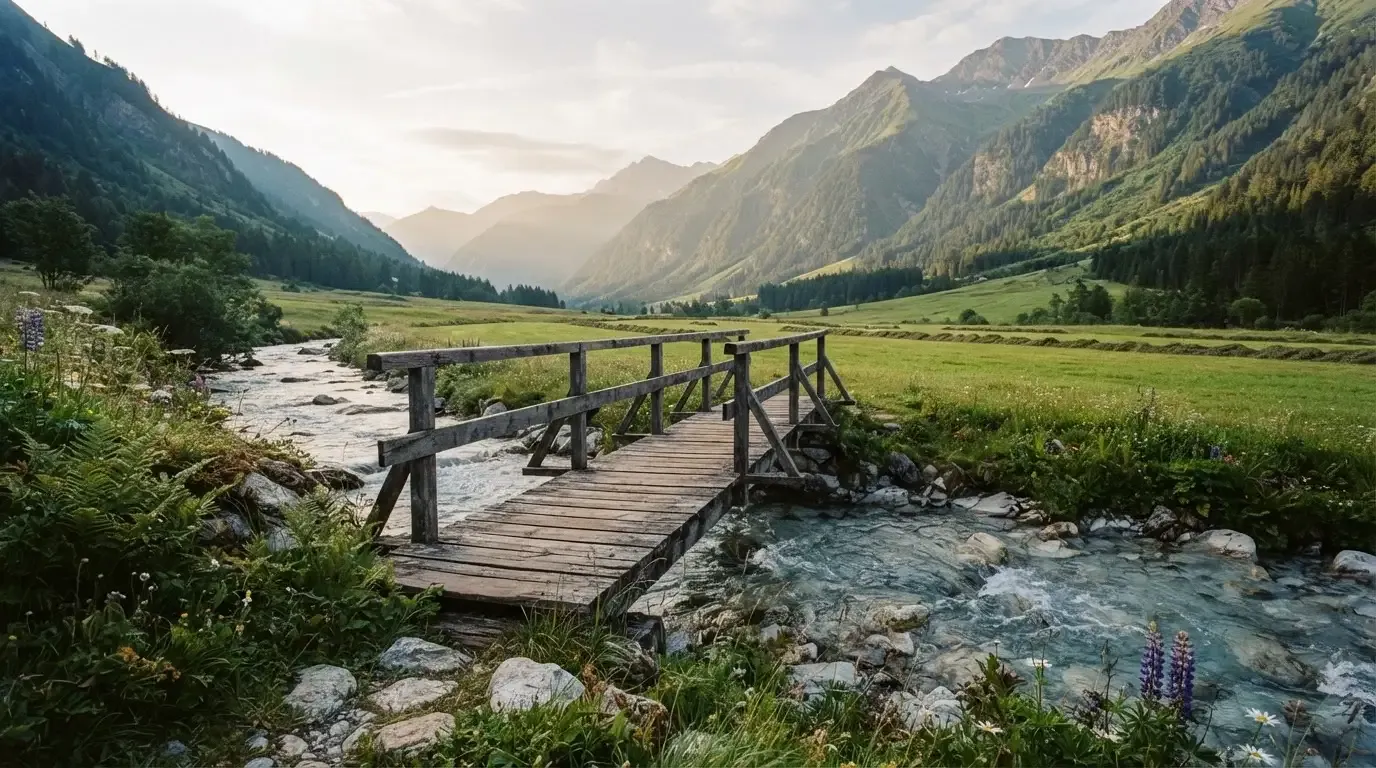 Ein Holzsteg führt über einen klaren Bergbach, im Hintergrund Wiesen und steile Hänge im Morgenlicht.