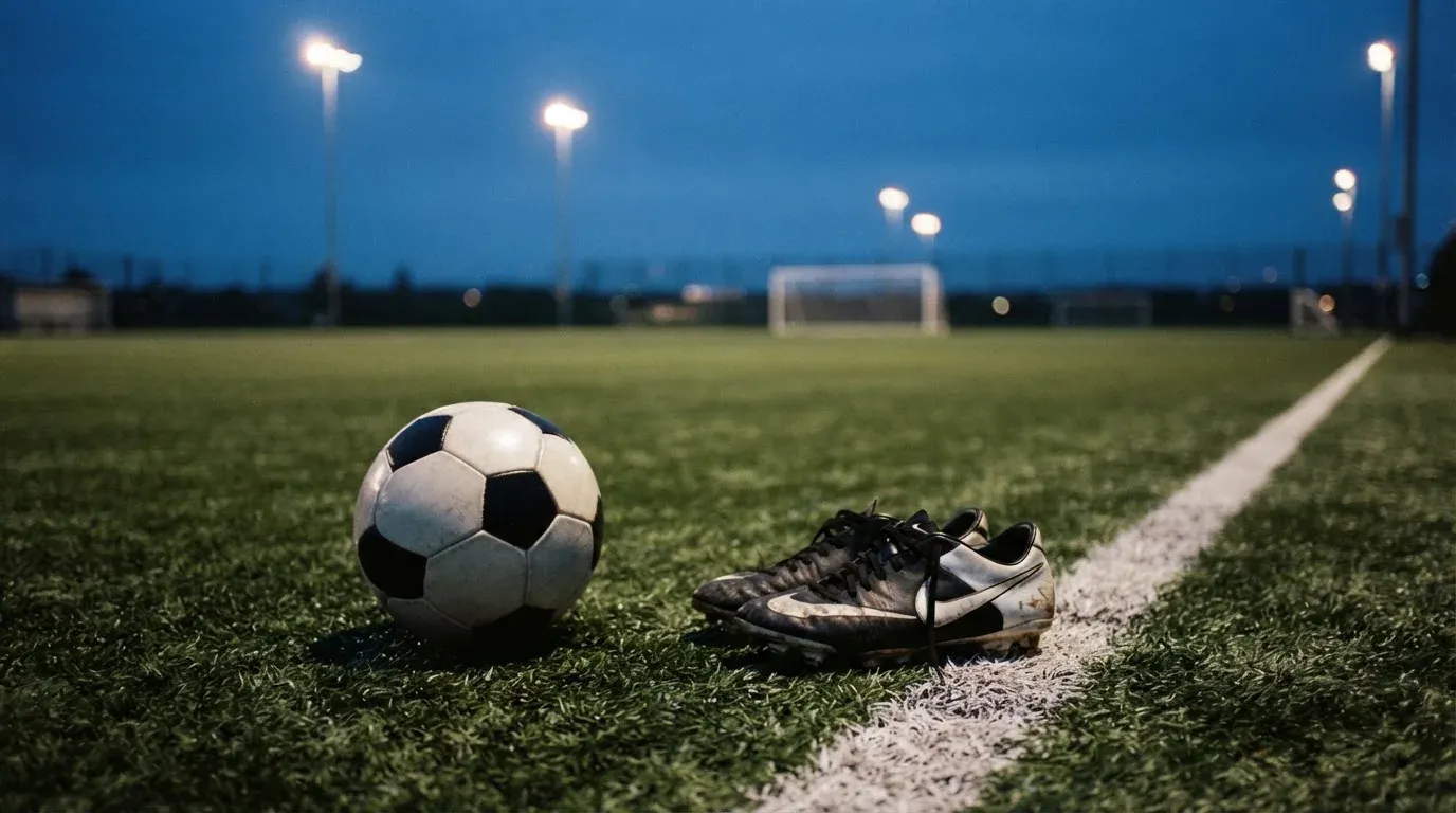 Ein Fussball und ein Paar Stollenschuhe stehen am Rand eines Kunstrasenplatzes, Flutlichtmasten ragen in den Abendhimmel.