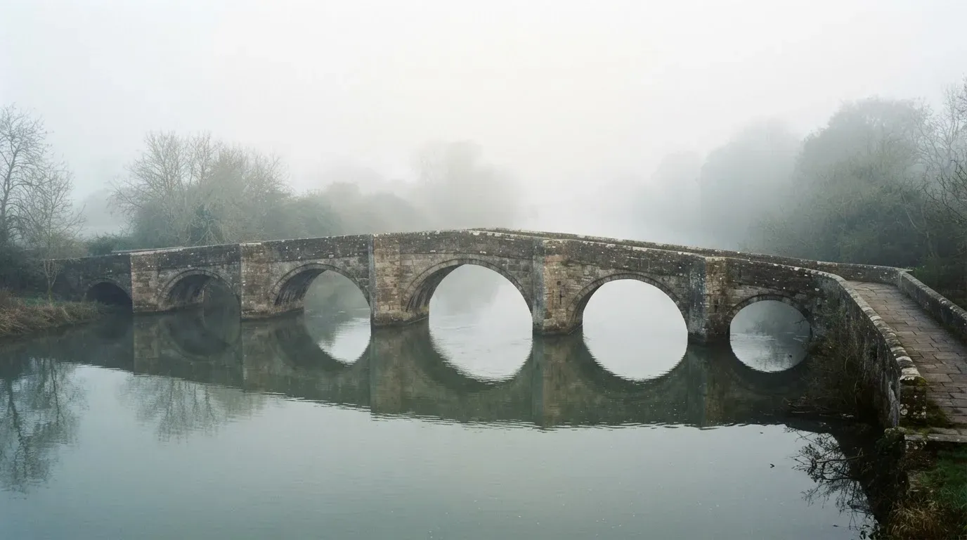Eine Steinbrücke führt im Frühnebel über einen Fluss, auf dem Fussweg ist niemand zu sehen.