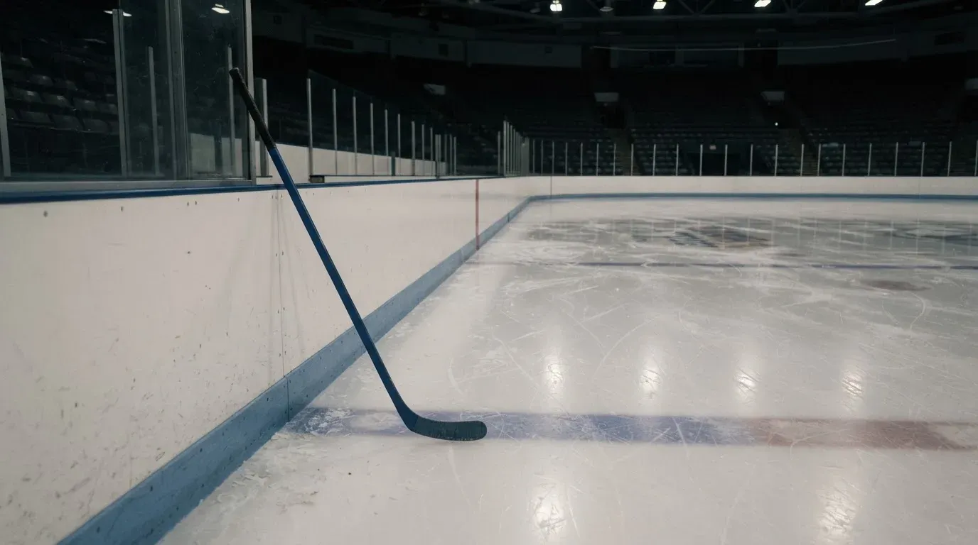 Ein Eishockeyschläger lehnt an der Bande einer leeren Halle, auf dem Eis spiegeln sich die Deckenlampen.