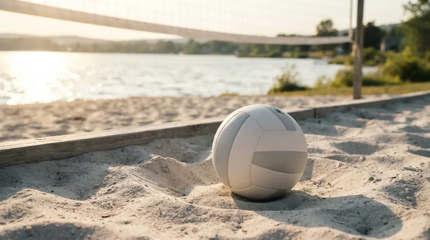 Ein Volleyball liegt auf dem Sand eines Strandfeldes am Seeufer, im Hintergrund glitzert das Wasser.