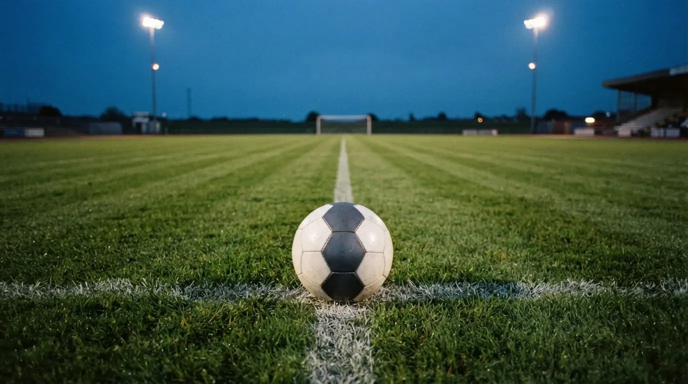 Ein Fussball liegt auf dem Mittelpunkt eines leeren Rasenplatzes, im Hintergrund ragen Flutlichtmasten in die Abenddämmerung
