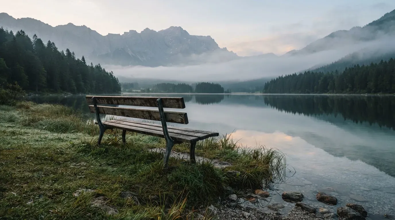 Eine Holzbank am Ufer eines ruhigen Bergsees, am gegenüberliegenden Ufer steigen dunkle Hänge in den Frühnebel