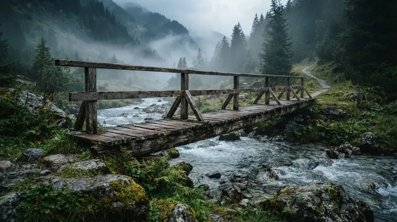Eine alte Holzbrücke über einen Gebirgsbach, Moos auf den Steinen, Nebelschwaden hängen im Tal