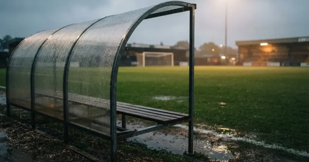Eine leere Ersatzbank unter einem Plexiglasdach am Spielfeldrand, Regentropfen auf der Scheibe, dahinter ein Rasenplatz