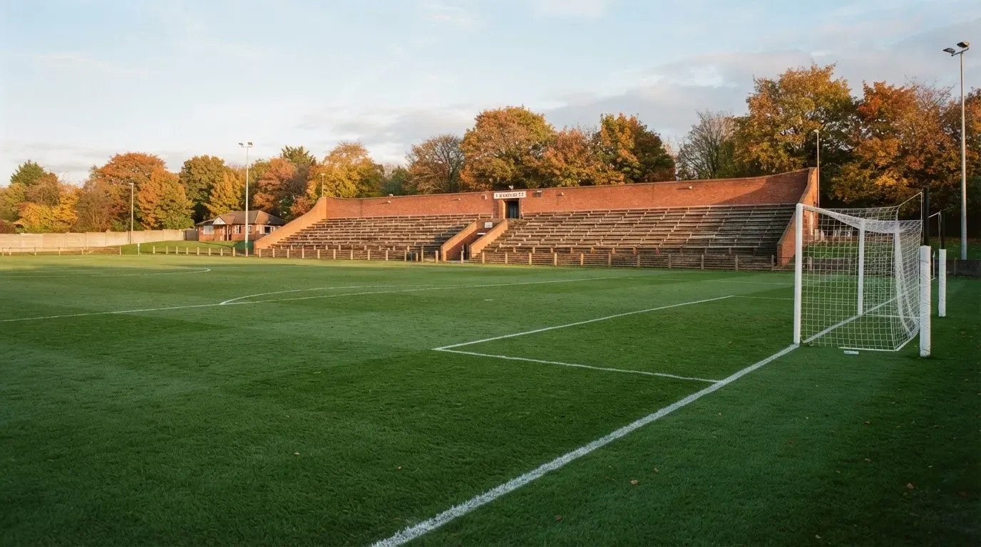 Leere Zuschauerränge eines kleinen Stadions im Herbstlicht, davor ein Rasenplatz mit frisch gezogenen Linien.