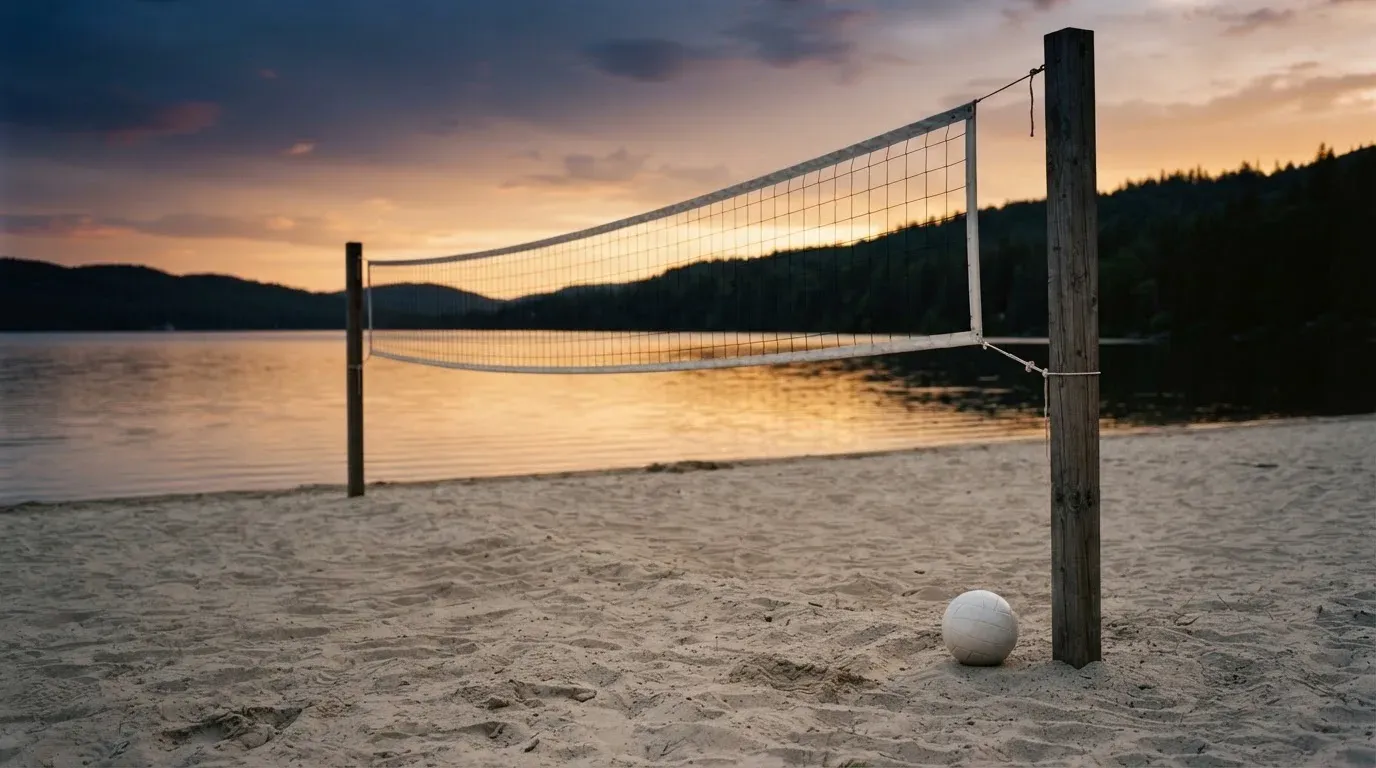 Ein Volleyballnetz steht an einem Seeufer in der Abenddämmerung, der Sand ist leer und das Wasser glatt.