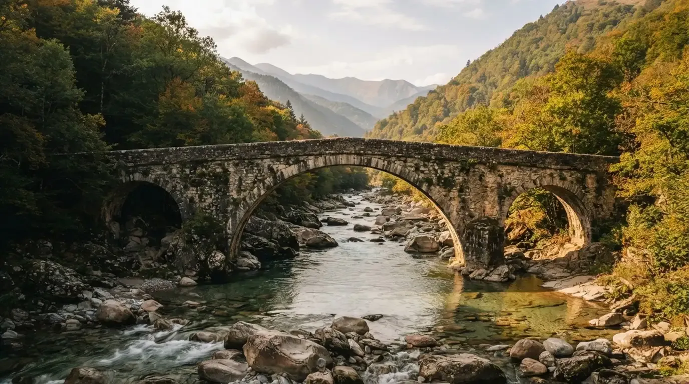 Eine alte Steinbrücke überspannt einen Gebirgsfluss, im Hintergrund steigen bewaldete Hänge an.