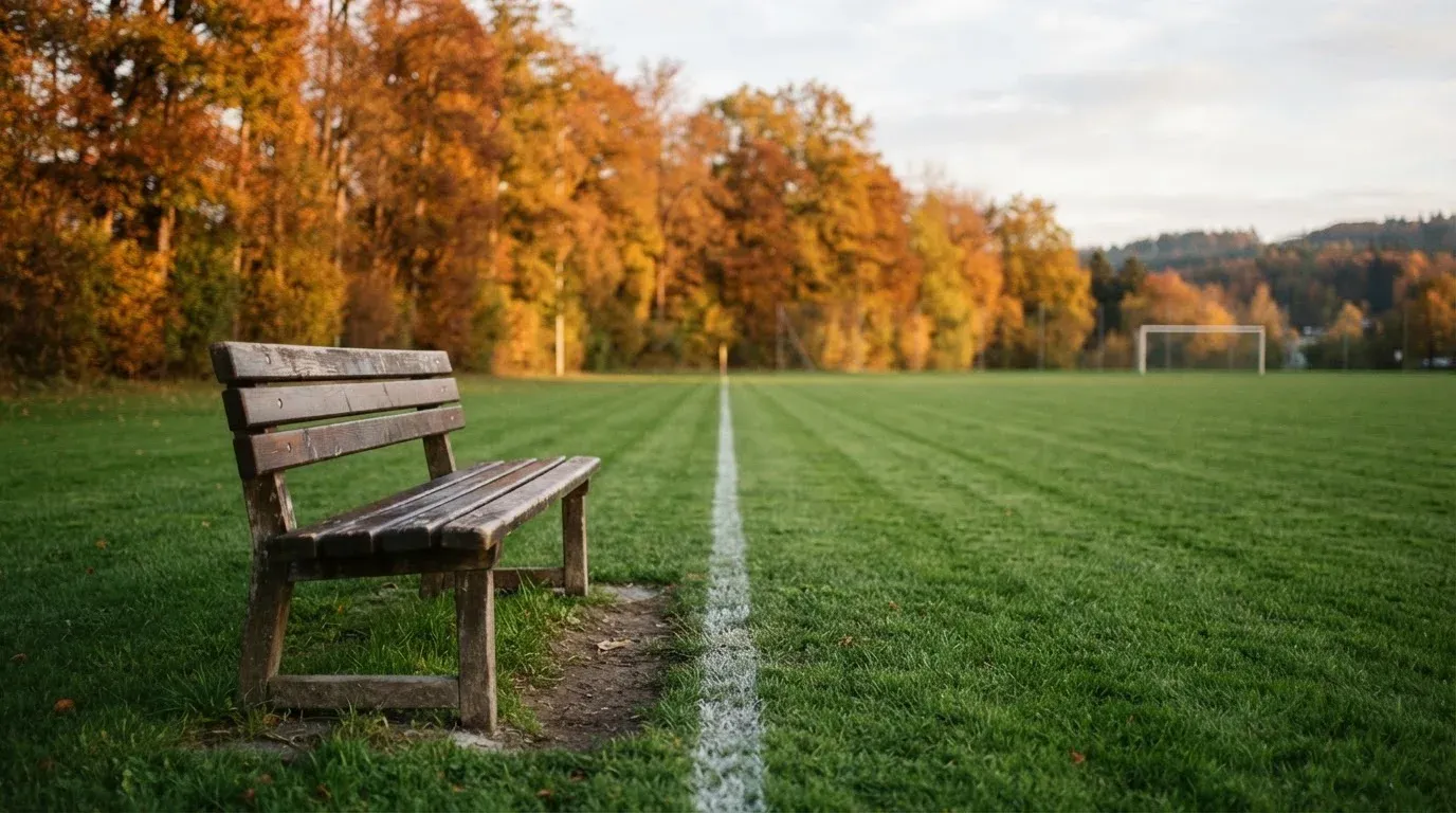 Eine hölzerne Bank steht an der Seitenlinie eines leeren Sportplatzes, dahinter Bäume im Herbstlicht.