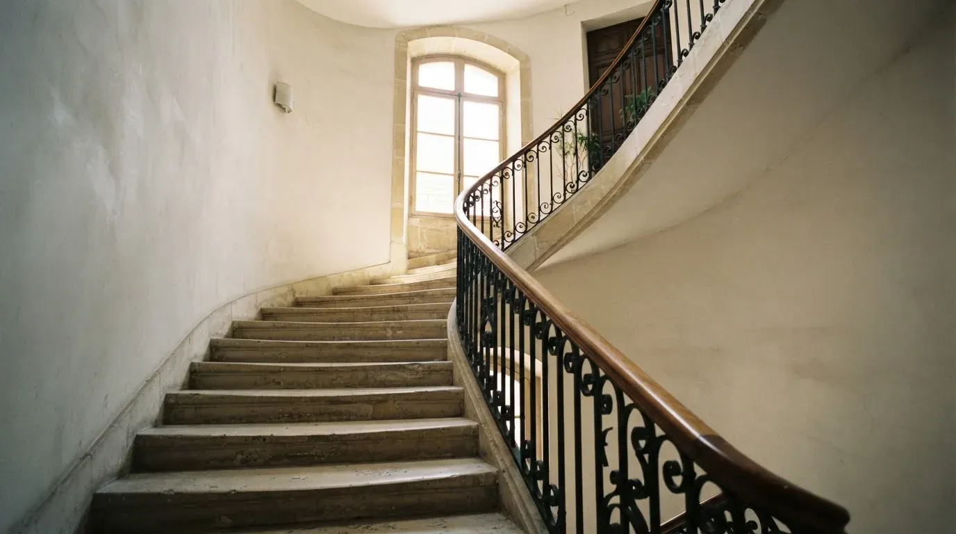 Ein Treppenhaus aus hellem Stein führt in einem Altbau nach oben, Licht fällt durch ein hohes Fenster.